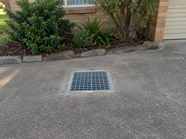 Square outdoor drain grate set in a concrete driveway beside garden beds in Belrose