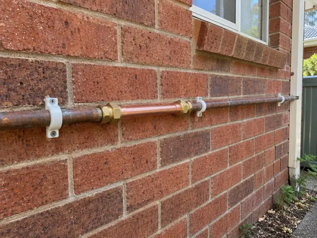 Repaired copper water pipe on a red brick exterior wall beside a window in Davidson